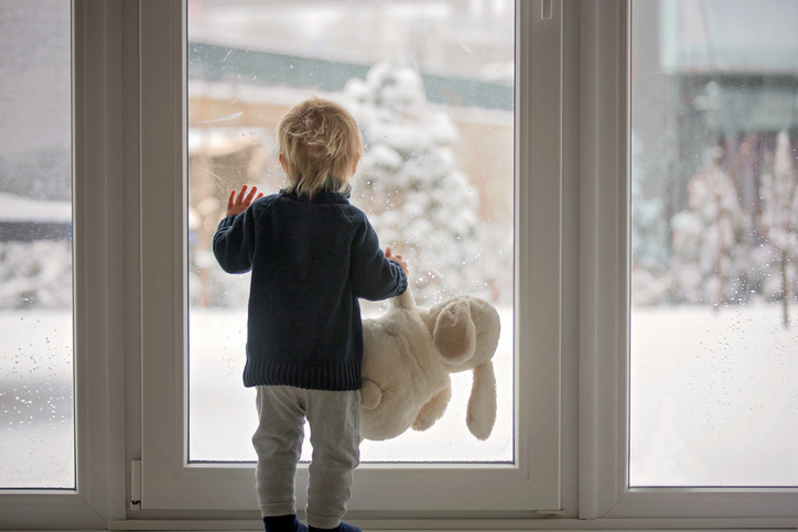 Toddler child standing in front of a big french doors, leaning against it looking outside at a snowy nature